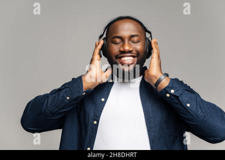 Studio portrait of relaxed cheerful African American man wearing and holding wireless headphones isolated on gray wall, enjoying listening to favorite music soundtrack, wellbeing, meditation concept Stock Photo