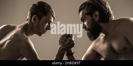Heavily muscled bearded man arm wrestling a puny weak man. Arms wrestling thin hand, big strong arm in studio. Two man's hands clasped arm wrestling