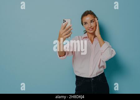 Elegant young lady in casual wear trying to take selfie while holding smartphone, isolated on blue Stock Photo