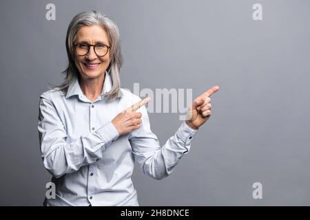 Successful senior mature gray-haired businesswoman in formal wear points fingers aside at empty copyspace, elder female entrepreneur ceo manager advertising, presenting, promoting isolated on gray Stock Photo