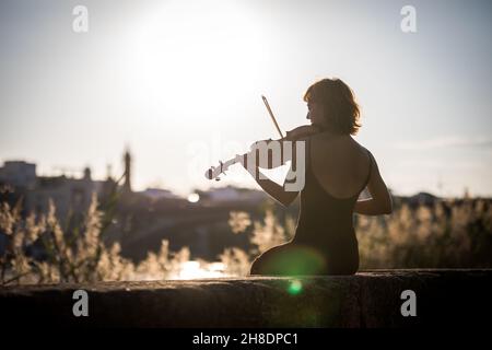 Young woman violinist posing playing on the street during sunset against light Stock Photo