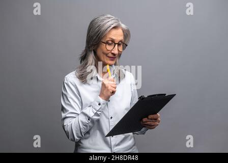 Modern elderly senior woman in formal wear taking notes standing with clipboard isolated on gray, mature female office employee writing down plans and ideas, checking up tasks Stock Photo
