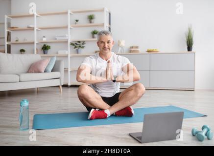 Sporty mature man practicing yoga online with instructor, sitting in lotus position in front of laptop in living room Stock Photo