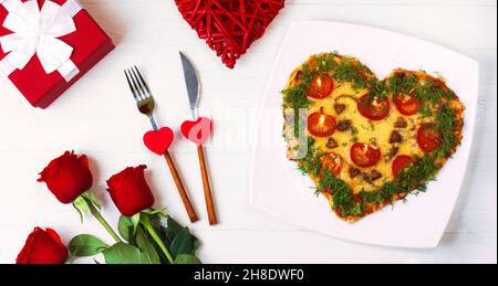Valentines Day food. Heart shaped pizza with greens, on a white wooden background. Red roses next to a gift in a red box. Cutlery with hearts. Top vie Stock Photo