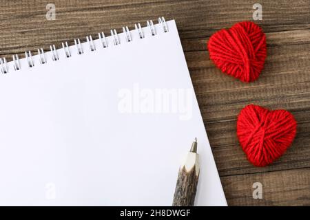 White sheet of notebook next to creative red handmade hearts, made of woolen threads, and wooden pen, on a natural wooden eco-friendly background. Val Stock Photo