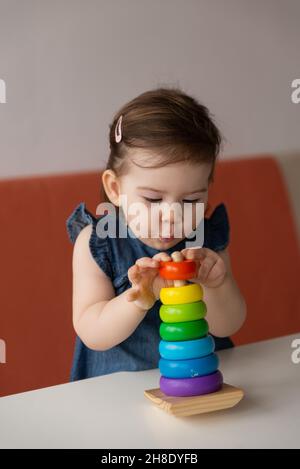 Pretty cute baby girl wearing jeans dress playing colourful toy wooden pyramid and making shooting emotional face. Little kid interesting explored and Stock Photo