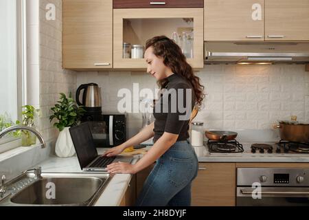 Smiling young woman cooking breakfast when having online conference with her colleagues Stock Photo