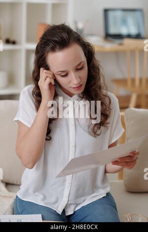 Pensive young female entrepreneur reading document when sitting on sofa at home Stock Photo