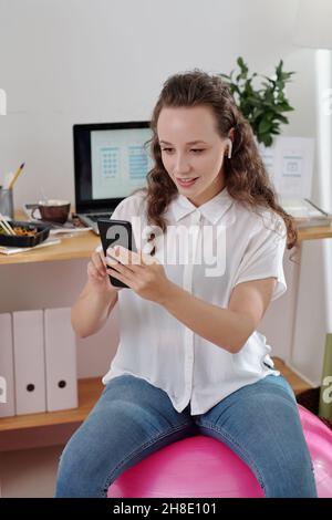 Pretty smiling young businesswoman sitting on fitness ball in her home office and video calling her colleague to discuss project Stock Photo