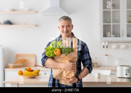 Mature man holding paper bag full of groceries from the supermarket, standing in kitchen interior and smiling Stock Photo