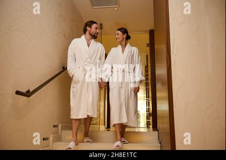 Adorable newlywed couple in love, dressed in white bathrobes, enjoying relaxation together in luxury spa during their honeymoon, holding hands as they Stock Photo