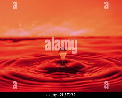 A red drop drips into the water and creates splashes of different shapes, due to the splashes waves are created on the water, the concept of a liquid Stock Photo