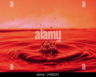 A red drop drips into the water and creates splashes of different shapes, due to the splashes waves are created on the water, the concept of a liquid Stock Photo