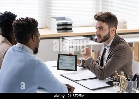 Bearded investment manager showing data on tablet to young couple while telling about benefits of cash investments Stock Photo