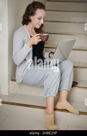Smiling young female entrepreneur sitting on steps at home, driking coffee and reading document on laptop screen Stock Photo