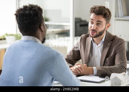 Confident young bearded recruiter sitting at table and asking candidate while holding job interview Stock Photo