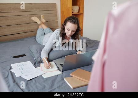Woman enjoying working from home, checking reports and agreements on laptop and taking notes in planner Stock Photo
