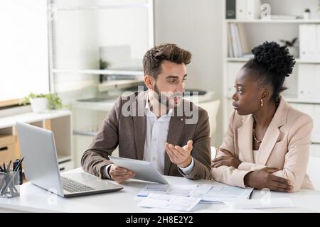 Young bearded manager using tablet while discussing income of company with colleague Stock Photo