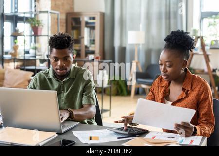 Concentrated young African American couple sitting at table in living room and calculating tax bills Stock Photo