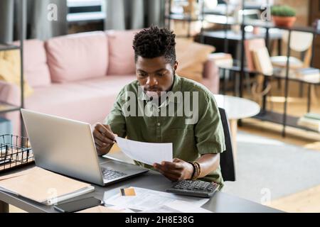Concentrated young African American taxpayer in green shirt sitting at table with laptop and calculator and checking tax form Stock Photo
