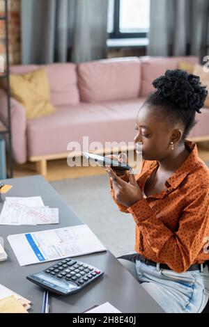 Serious young African American woman sitting at table with calculator and photographing tax document on phone Stock Photo
