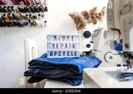 Reuse, repair, upcycle text on light board on sewing machines background. Stack of old jeans, Denim clothes, scissors, thread and sewing tools in Stock Photo