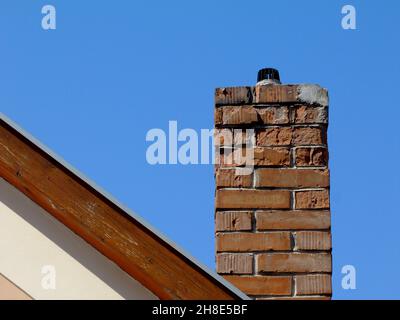 damaged clay brick chimney. spalled brick texture. wood trim. house gable end wall. sloped roof. blue sky. white stucco exterior. side elevation view Stock Photo