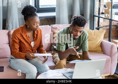 Serious young African American man sitting at coffee table with laptop and examining finances with girlfriend Stock Photo