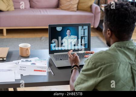 Over shoulder view of African American man sitting at table in living room and talking to financial consultant online Stock Photo