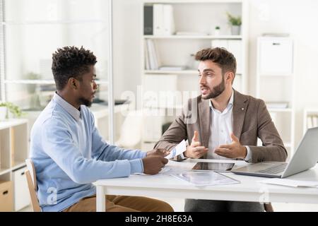 Handsome young businessman talking to African American accountant while preparing financial report with him Stock Photo