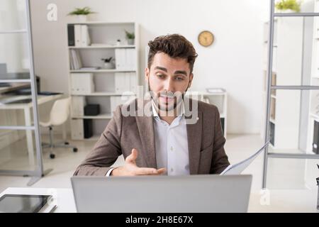 Confident young bearded businessman in jacket gestuing hand and commenting papers while holding online meeting using laptop Stock Photo