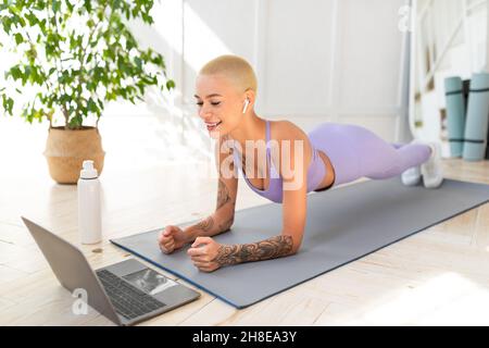 Workout for weight loss and keep fit. Happy young woman in sportswear doing plank on mat and looking at laptop Stock Photo