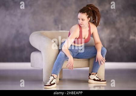 Woman with box braids dressed in tank top and jeans sits in chair thinking. Stock Photo
