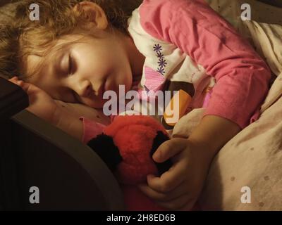 Adorable little girl asleep in her bed dimly lit by a bedside table lamp with a warm bulb Stock Photo