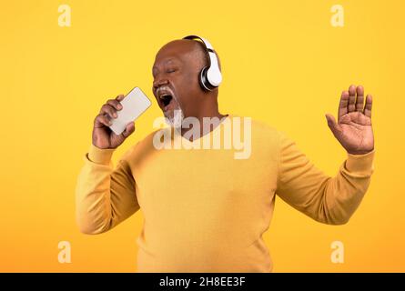 Cheerful senior black man listening to music in wireless headphones, singing, using smartphone as microphone, mockup Stock Photo