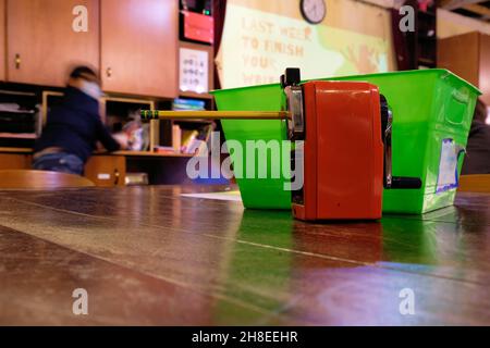 A pencil sharpener with a pencil inserted for sharpening on a wooden table in a classroom work station with a blurred motion student in background. Stock Photo