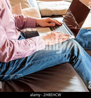 Side view of crop anonymous businessman sitting on sofa and typing on laptop while working at home Stock Photo