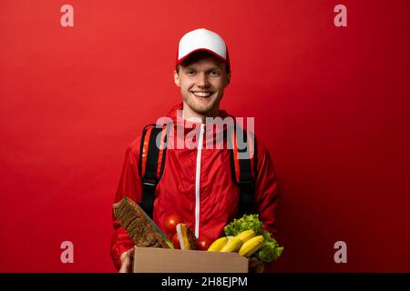 Delivery man in a red uniform with a thermal bag on a shoulder delivering food to a customer at home. Online grocery shopping service concept.