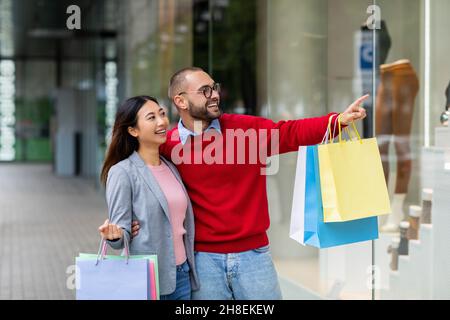 Affectionate young interracial couple standing on street with shopping bags, pointing at store window near city mall Stock Photo