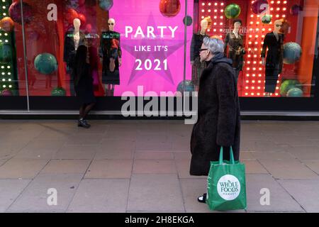 As the new Covid-19 variant known as Omicron becomes more prevalent in the UK, a shopper carrying a bag from a Wholefoods Market, walks past an ad outside John Lewis on Oxford Street, where a window tells consumers to party like it's 2021, on 29th November 2021, in London, England. Stock Photo