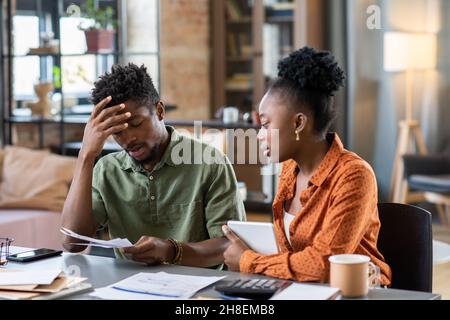 Puzzled young blackman touching forehead and looking at bills while his girlfriend supporting him Stock Photo