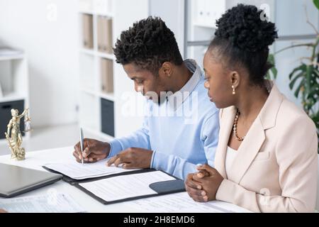 Serious young Blackman sitting at table in lawyers office and signing divorce papers Stock Photo
