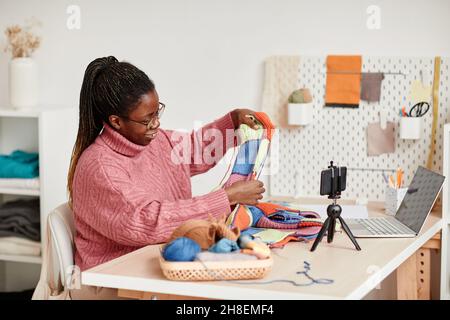 Portrait of smiling African-American woman knitting at home and recording video or livestream, copy space Stock Photo