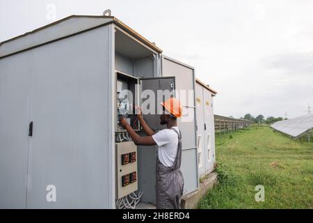 African american electrician in uniform and helmet standing near box with cables and doing service work on solar station. Maintenance, people and green energy concept. Stock Photo