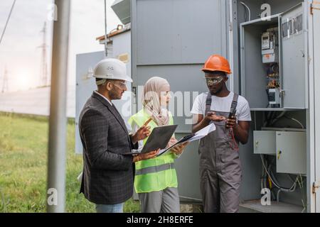 Indian man and muslim woman with clipboard and laptop talking with african american technician at solar station. Multiracial people examining process of green energy production. Stock Photo