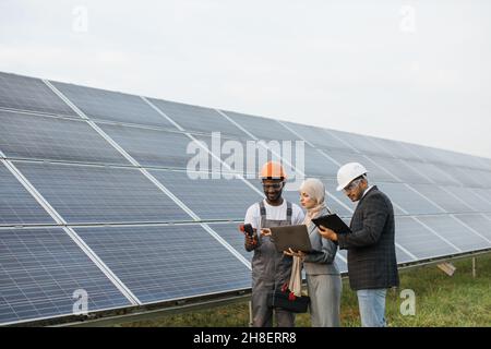 Muslim woman and indian man standing with african american technician outdoors and checking temperature of solar panels. Inspectors using laptop and clipboard, worker holding thermal imager. Stock Photo