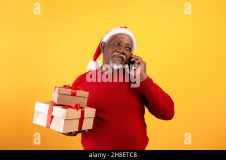 Senior black man holding Xmas gift boxes, wearing Santa hat, speaking on smartphone on orange studio background Stock Photo