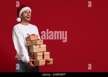 Holiday Ad. Cheerful Young Man Wearing Santa Hat Holding Stack Of Gifts Stock Photo
