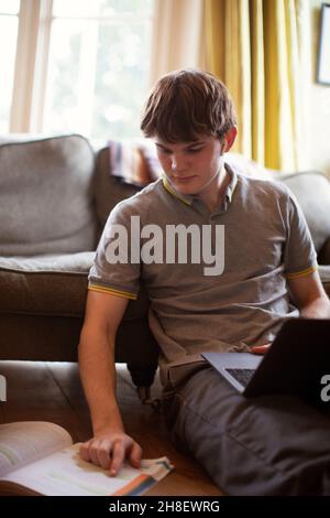 Focused teenage boy with laptop and textbook doing homework Stock Photo