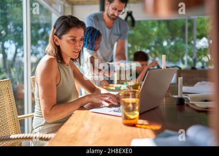 Woman working at laptop in dining room with family Stock Photo